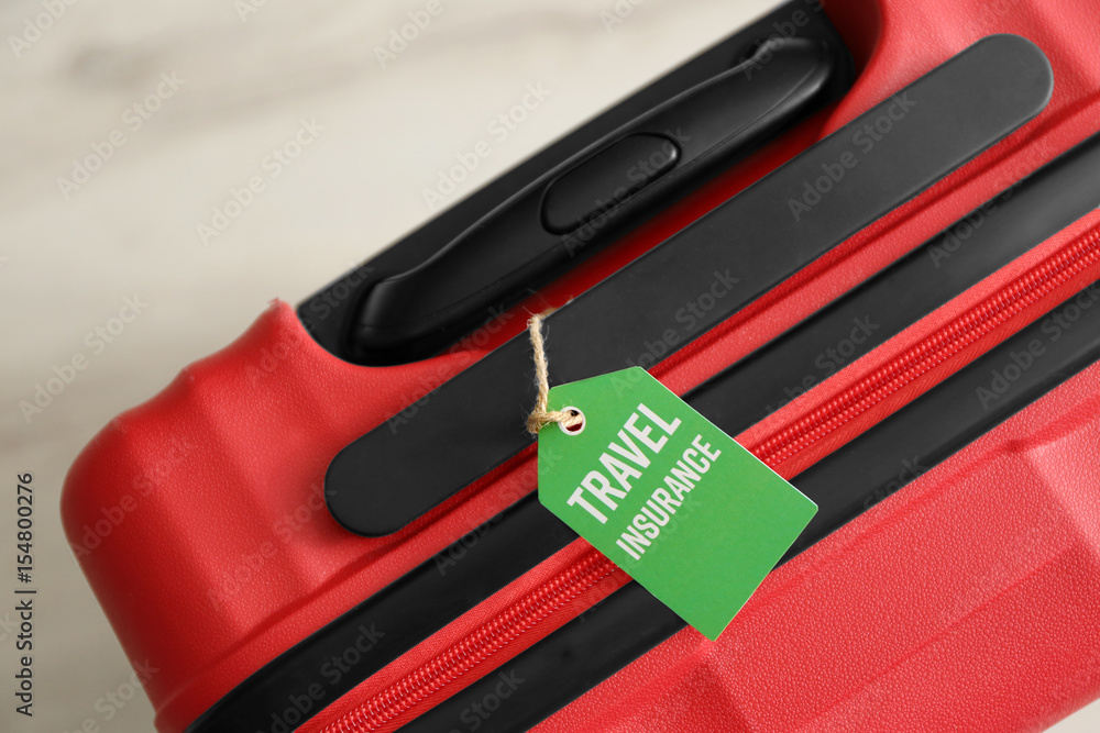 Red suitcase with label, closeup. TRAVEL INSURANCE concept