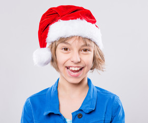 Wall Mural - Emotional portrait of attractive caucasian little girl wearing Santa Claus red hat. Funny cute smiling child 10 year old looking at camera on gray background. Winter holiday christmas concept. 