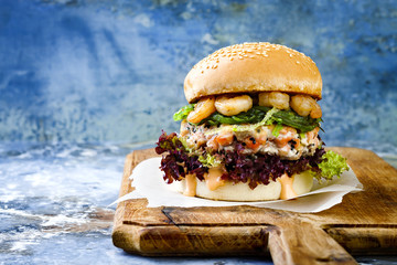 Asian style salmon burger with grilled shrimps, seaweed, lettuce and spicy sriracha mayo sauce served on pieces of brown paper on a rustic wooden board. Bright blue background with copyspace