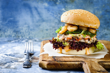 Wall Mural - Asian style salmon burger with grilled shrimps, seaweed, lettuce and spicy sriracha mayo sauce served on pieces of brown paper on a rustic wooden board. Bright blue background with copyspace