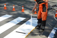 Worker Is Painting A Pedestrian Crosswalk. Technical Road Man Worker Painting And Remarking Pedestrian Crossing Lines On Asphalt Surface Using Paint Sprayer Gun.