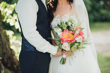 The bride in a white dress and veil and the groom in a shirt and vest are standing on a green glade and holding a wedding bouquet of flowers and greens