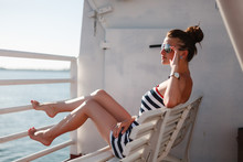 Young Woman In A Striped Dress And Goggles From The Sun Sitting On A Bench On Board Ship Or Ferry And Looking Into The Distance, Enjoying The Warm Breeze From The Sea