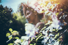 True Tilt-shift View Of Young Cute Black Female Teenager With Chamomile Wreath On Her Head With Curly Afro Hair, Standing In Beautiful Summer Garden Surrounded By Light Pink And Light Blue Flowers