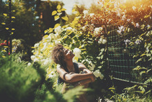 Cute Young Curly Afro American Girl Enjoying Sun While Sitting On The Ground In Private Garden, Happy Black Teenage Female Spending Sunny Morning In Park With Beautiful Flowers Near Fence