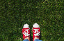 Legs In Old Red Sneakers On Green Grass. View From Above. The Concept Of Youth, Spring And Freedom