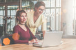 © foxyburrow - Two businesswomen working together. On desk is laptop. Woman is standing near table, girl is sitting next to her.