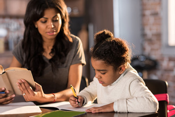 mother looking at cute little daughter doing homework at home, homework help concept