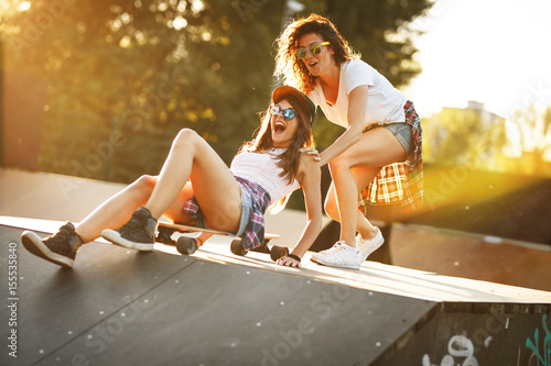 Zdjęcie XXL Dwie koleżanki, grając z deskorolka w skate park. Jedna dziewczyna pchanie innych od tyłu. Śmiać się i zabawy.