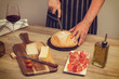 Hombre cortadndo un pan redodno y casero junto a una copa de vino tinto, jamón, queso, pan sobre una emsa de madera rústica. Vista superior