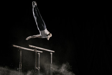 Male athlete performing difficult exercise on gymnastic parallel bars with talcum powder. Isolated on black.