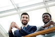 Low angle portrait of handsome bearded businessman with African-American colleague looking down from glass balcony and smiling holding cups during break