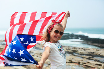 American flag. Little smiling  patriotic girl with long blond hair, red head band  bandana and sunglasses holding an american flag waving in the wind on the ocean beach. National 4 july. Memorial day