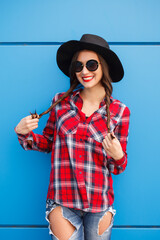 Wall Mural - Portrait of beauty fashion smiling hipster girl in hat and sunglasses on blue background