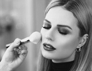 Process of making makeup. Mak-up artist working with brush on model face. Portrait of young blonde woman in beauty saloon interior. Applying tone to skin.