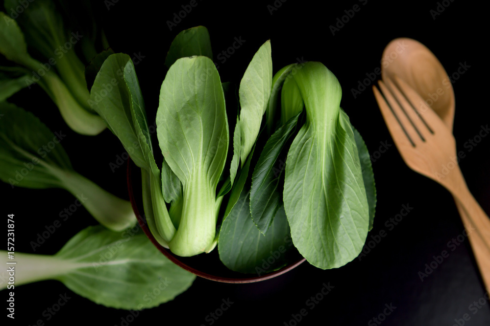 close up Fresh baby green bok choy in bowl with woodden spoon and fork on the  black background , overhead or top view shot - obrazy, fototapety, plakaty 
