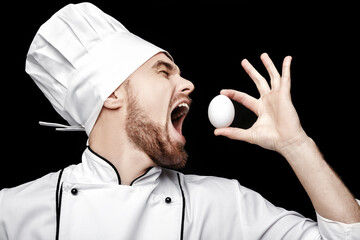 Wall Mural - Young bearded man chef In white uniform holds  egg on black background