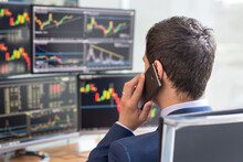 Over The Shoulder View Of And Stock Broker Trading Online While Accepting Orders By Phone. Multiple Computer Screens Ful Of Charts And Data Analyses In Background.