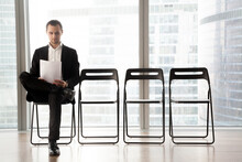 Confident Job Applicant Reads Resume While Sitting On Chair In Row In Office And Waiting His Turn On Interview. Young Guy Wearing Suit Prepares For Recruitment Meeting With Employer In Waiting Room