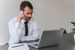 © bnenin - Portrait of a elder businessman with beard speaking on his smartphone while working on a computer at his desk. He is in a office his notebook in front of him like an insurance or bank manager.