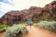 Happy Senior Woman Hiking In A Red Rock Sandstone Canyon. The Active, Retired Woman Is Enjoying A Walk Along A Scenic Trail With Vibrant Red Rock Cliffs In The Background.