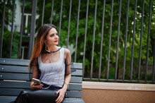 Fashionable Woman Look At White Shirt, Black Transparent Clothes, Leather Pants, Posing At Street And Sitting On Bench With Cell Phone And Headphones. Fashion Girl Listening Music On Mobile.