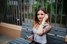 Fashionable Woman Look At White Shirt, Black Transparent Clothes, Leather Pants, Posing At Street And Sitting On Bench With Cell Phone And Headphones. Fashion Girl Listening Music On Mobile.