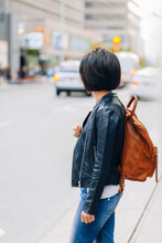 Portrait Of Young Girl Woman With Short Dark Hair Bob Style, In Blue Jeans And Leather Biker Jacket, With Yellow Brown Backpack On Shoulder, Standing In City Street, View From Back Behind