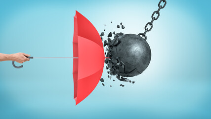 a male hand holding an open red umbrella which protects from a collision with a broken wrecking ball