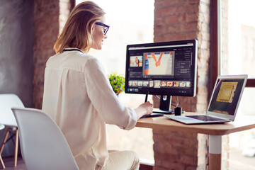 Smilimg blond girl is working at home on computer, using her laptop also. She is a graphic designer. Retouch is her hobby, she is self employed freelancer