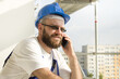 Uśmiechnięty pracownik budowlany w stroju roboczym i w kasku na głowie rozmawia przez telefon. Praca na dużej wysokości. Rusztowania w tle.