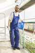 Pracownik budowlany w stroju roboczym, rękawicach ochronnych trzyma kask i młotek. Praca na dużej wysokości. Rusztowania w tle.