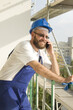 Uśmiechnięty pracownik budowlany w stroju roboczym i w kasku na głowie rozmawia przez telefon. Praca na dużej wysokości. Rusztowania w tle.