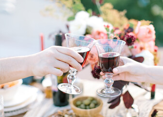 Close -up Of Couple Hands Toasting Red Wine on festively table with flowers and fresh food background. birthday, event, wedding concept. Man and woman drinking red wine.