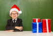 © Ermolaev Alexandr - Happy boy in red christmas hat with gift boxes near empty green blackboard