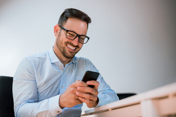 Portrait of a happy smiling businessman in eyeglasses using smartphone while sitting at the office