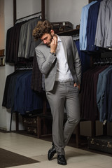 Wall Mural - Full length portrait of bearded man in suit standing with crossed arms near the rack with suits in a shop while looking at the camera