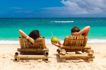 Beach summer couple on island vacation holiday relax in the sun on their deck chairs on the tropical beach. Idyllic travel background.