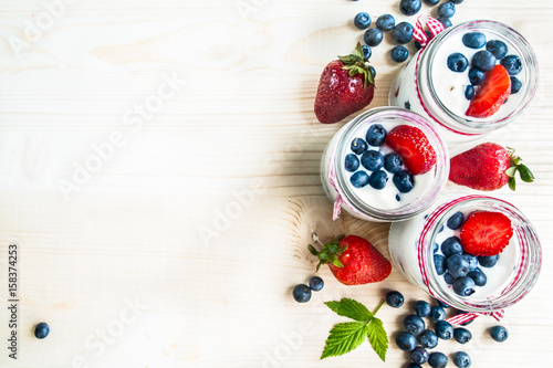 Zdjęcie XXL Trzy słoiki jogurtu z jagodami i truskawkami, kwadrat. Jagody jagód i truskawek są rozrzucone na stole. Gotowanie, zdrowa żywność, pakowanie żywności