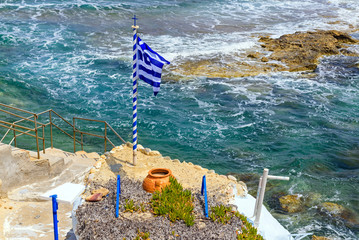 Greek national flag over seascape, beautiful turquoise Aegean Sea. Mandrakia village, Milos Island. Greece.