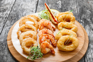 Seafood platter with deep fried squid rings, shrimp and onion rings decorated with lemon on cutting board on wooden background. Mediterranean appetizers. Top view