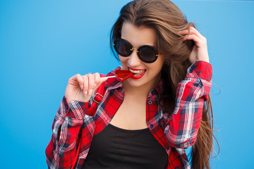 Fashion portrait of pretty smiling woman in sunglasses with lollipop against the colorful blue wall. outdoor