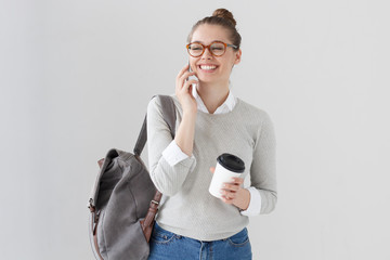 Wall Mural - Attractive stylish girl laughing cheerfully while talking on mobile phone to her boyfriend, isolated on gray background. Conversation concept. Happy hipster teacher wearing glasses with coffee