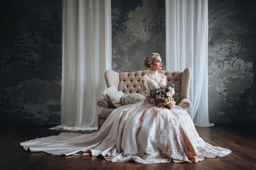 Beautiful bride in a cream wedding dress with a wedding bouquet, with orange and white flowers. Studio, gray background, modern, sofa, lace dress.
