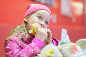 Wall Mural - Adorable blonde girl in pink jacket and hat eat burger in fast food restaurant