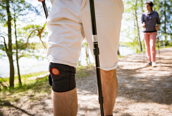 Man with bandage on knee. Rehabilitative sport in Finland - nordic walking. Man and mature woman hiking in green sunny forest. Active people outdoors. Scenic peaceful Finnish summer landscape.