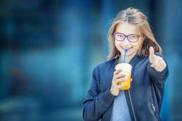 Cheerful pre-teenage girl drinking juice. Girl with dental teeth braces and glasses.