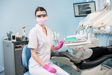 Portrait Of Attractive Female Dentist With Patient In The Dental Clinic. Doctor Wearing Glasses, Mask, Uniform And Pink Gloves. On The Background Monitor With X-ray The Patient's Teeth. Dentistry