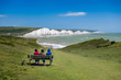 Three people sitting on a bench watching the waves on a windy but sunny day at Cuckmere Haven Sussex UK