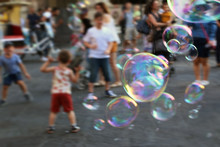  Soap Bubbles On The Town Square And Blurry Children  That Are Chasing Them In The Background